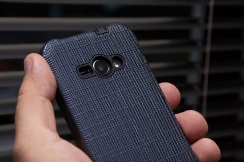 Tangan memegang smartphone dengan casing biru bertekstur di dekat jendela