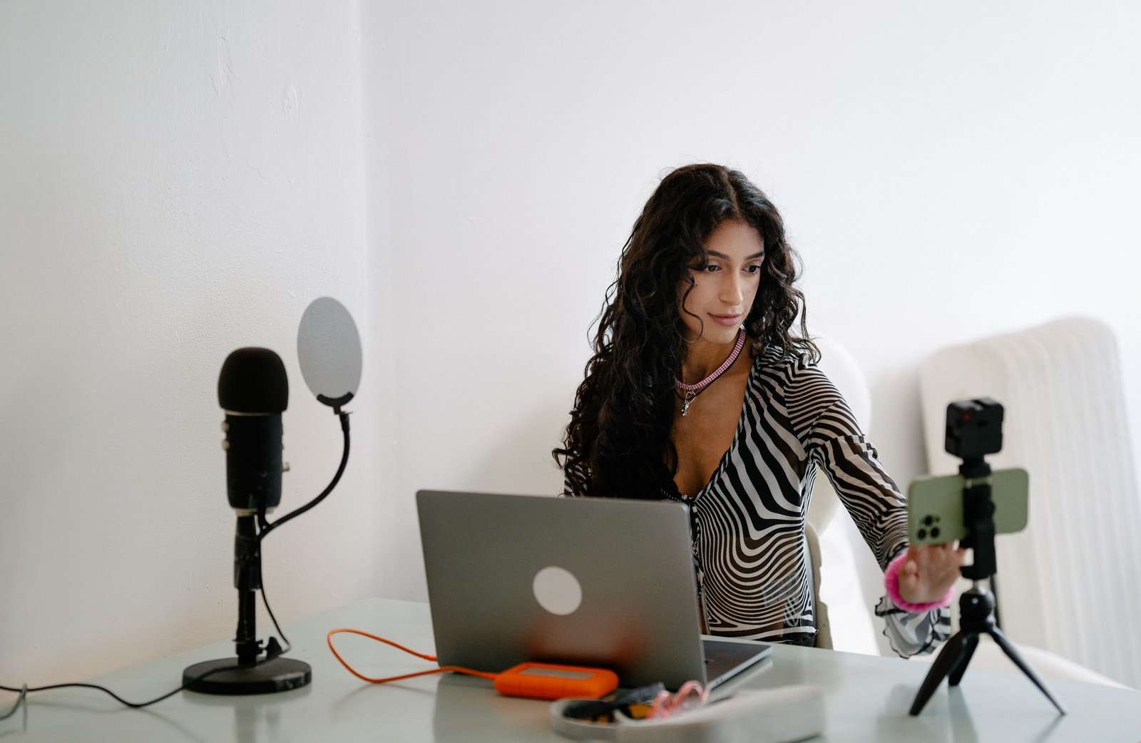 Perempuan menyiapkan siaran langsung dengan ponsel di tripod, laptop, dan mikrofon di atas meja