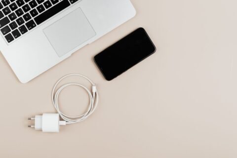 Ponsel, kabel charging, dan adaptor diletakkan di samping laptop pada permukaan meja berwarna krem.