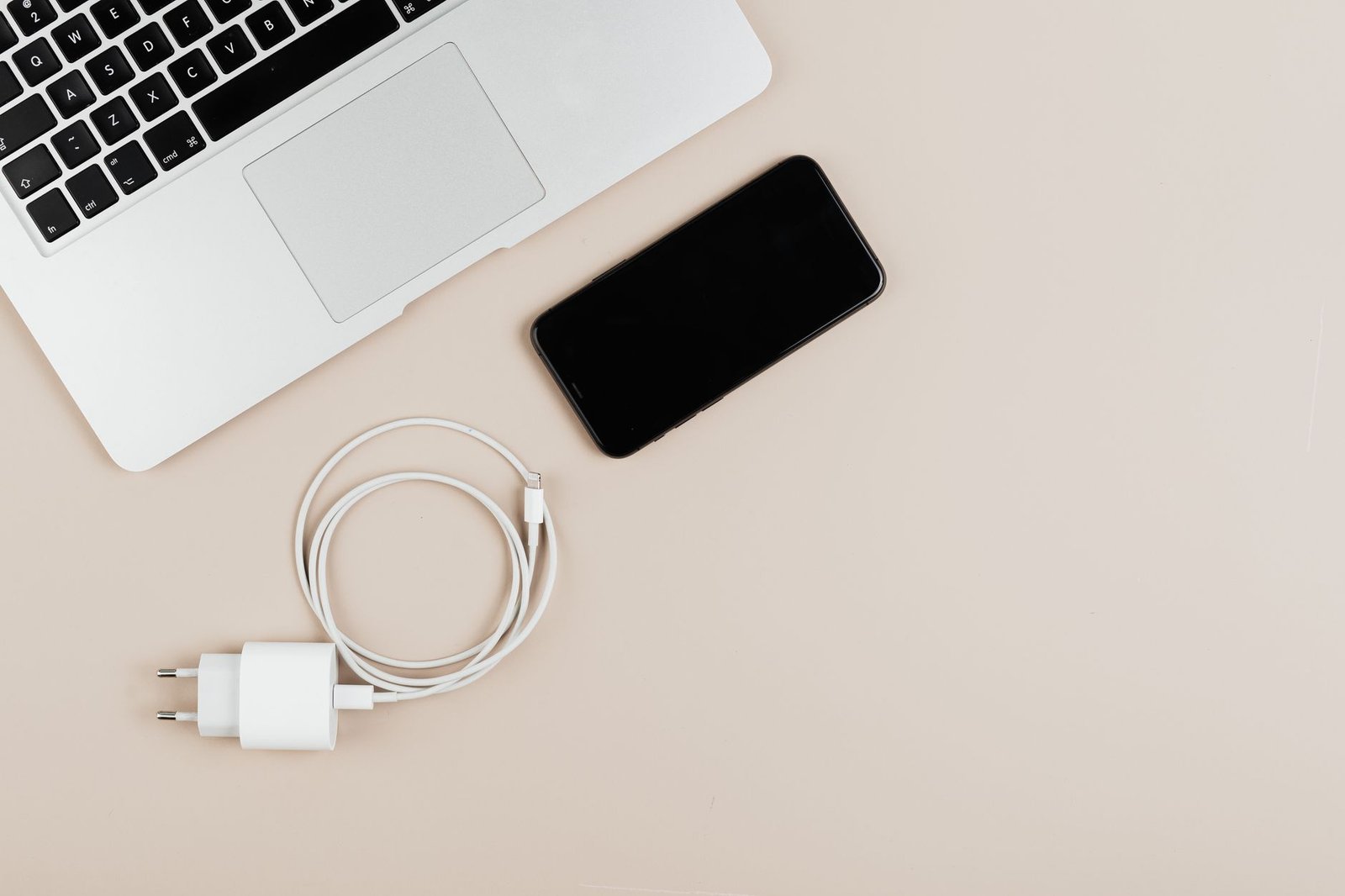 Ponsel, kabel charging, dan adaptor diletakkan di samping laptop pada permukaan meja berwarna krem.