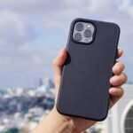 Tangan memegang iPhone bercasing hitam bertekstur di depan latar kota yang buram