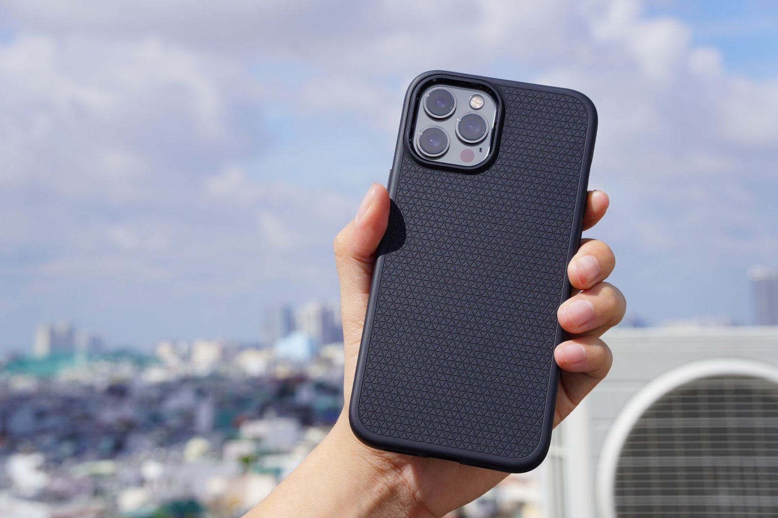 Tangan memegang iPhone bercasing hitam bertekstur di depan latar kota yang buram