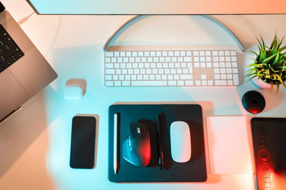 Tampilan atas meja kerja dengan smartphone, laptop, keyboard, mouse, earbud, dan aksesori teknologi lainnya.