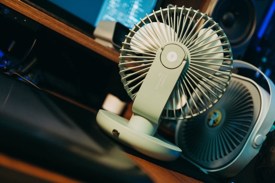 Kipas meja kecil berwarna hijau terlihat di atas meja kerja, dengan kipas lain dan perangkat audio di latar belakang.