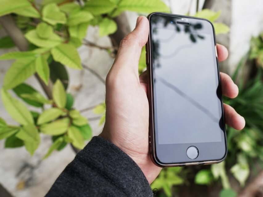 Seseorang memegang iPhone dengan layar kosong di luar ruangan, berlatar dedaunan hijau.