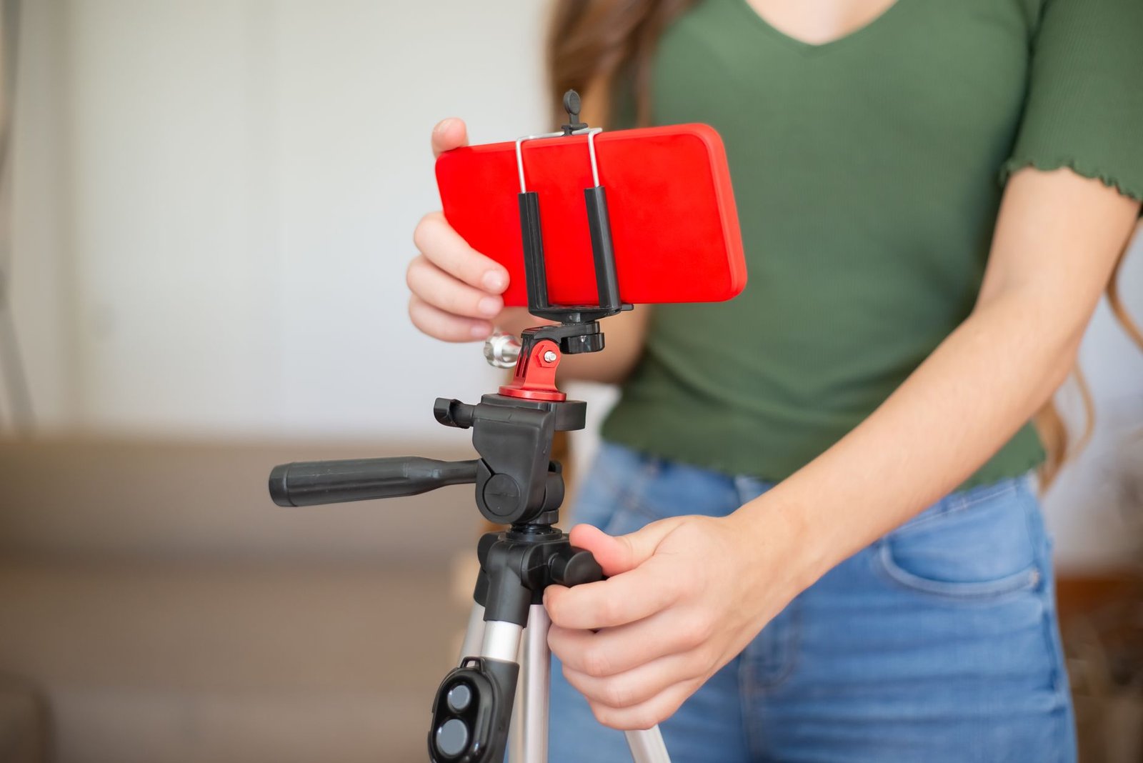 Seorang perempuan memasang smartphone merah pada holder di atas tripod untuk merekam video di dalam ruangan.