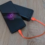 Ponsel sedang diisi daya dari power bank menggunakan kabel oranye di atas meja kayu