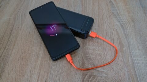 Ponsel sedang diisi daya dari power bank menggunakan kabel oranye di atas meja kayu