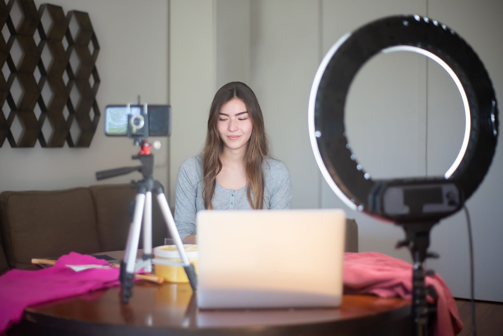 Perempuan membuat vlog di dalam ruangan dengan smartphone pada tripod, ring light, dan laptop di atas meja