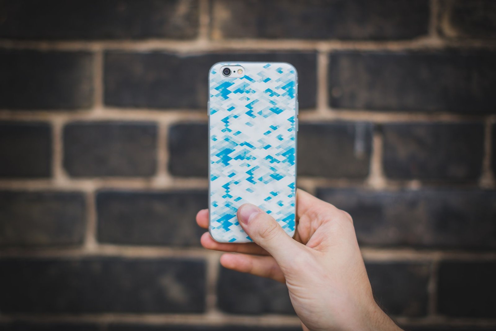Tangan memegang ponsel dengan casing bermotif biru-putih di depan dinding bata gelap