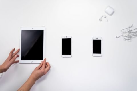 Tablet, dua ponsel, dan earphone putih tersusun di atas permukaan putih.