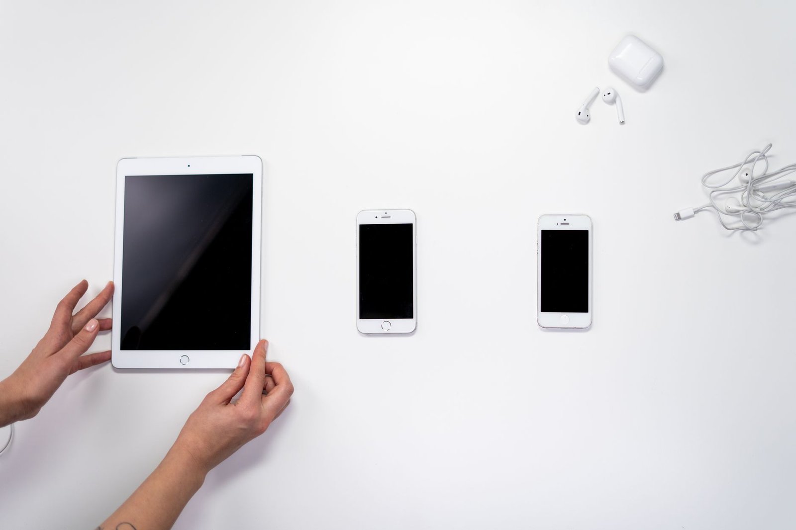 Tablet, dua ponsel, dan earphone putih tersusun di atas permukaan putih.