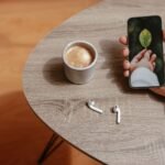 Seseorang memegang smartphone di atas meja kayu dengan secangkir kopi dan earbuds di dekatnya