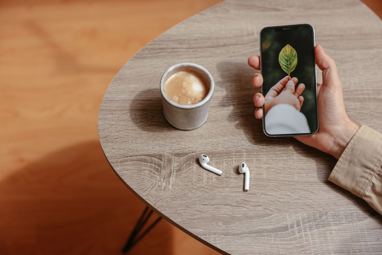 Seseorang memegang smartphone di atas meja kayu dengan secangkir kopi dan earbuds di dekatnya