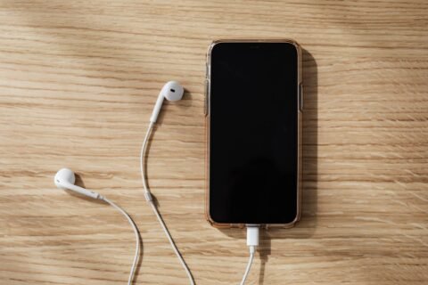 Ponsel dalam casing transparan sedang diisi daya dengan kabel, ditemani earphone putih di atas meja kayu.