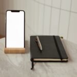 Smartphone berdiri pada holder kayu di atas meja marmer di samping buku catatan dan pena.