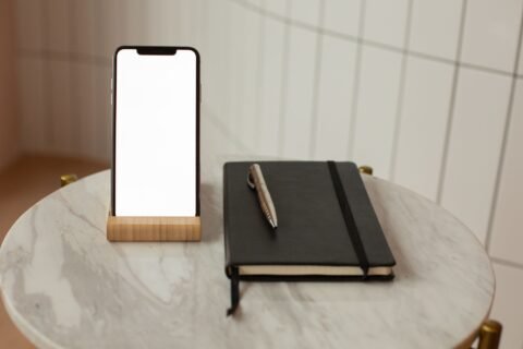 Smartphone berdiri pada holder kayu di atas meja marmer di samping buku catatan dan pena.