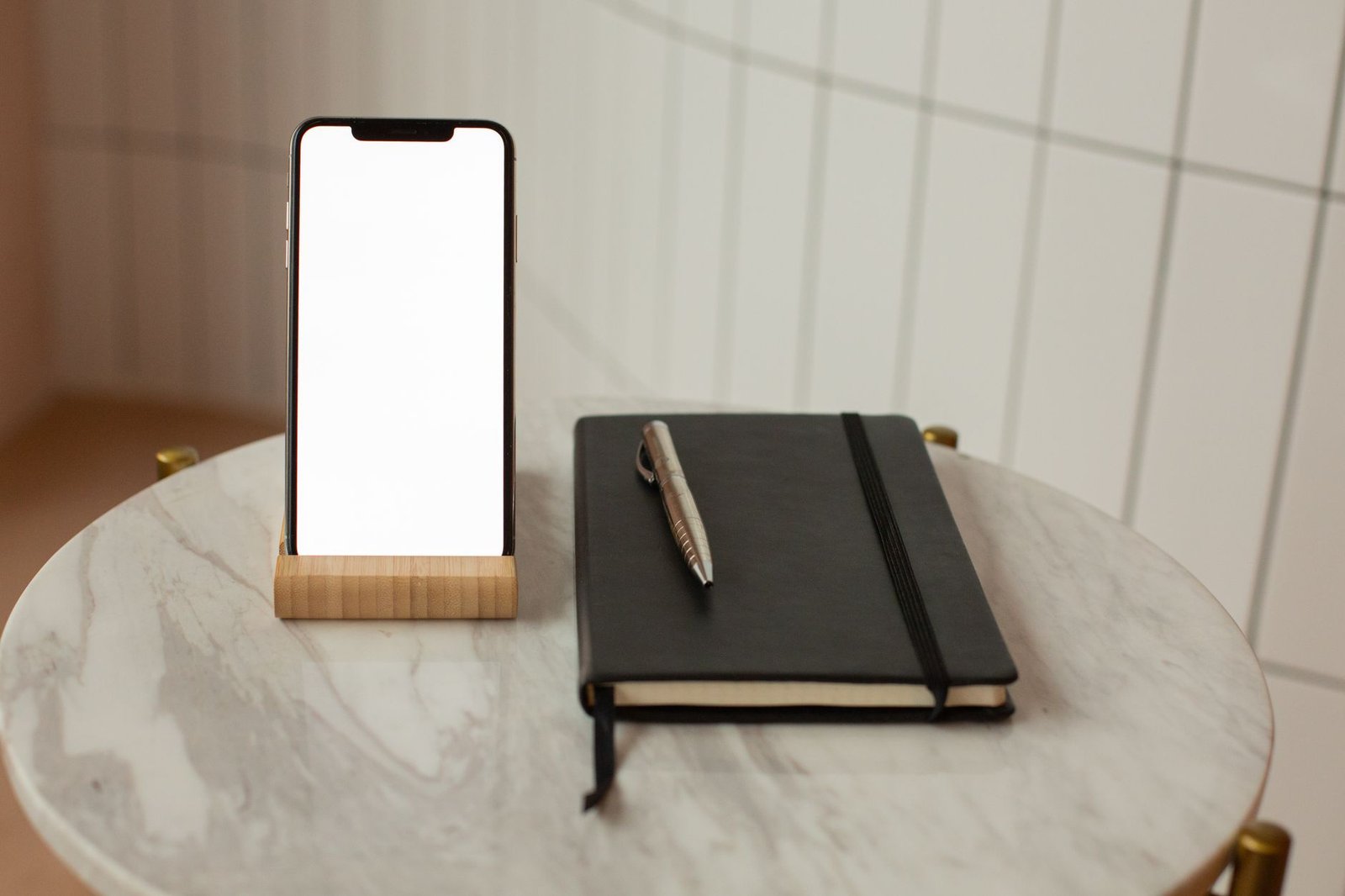 Smartphone berdiri pada holder kayu di atas meja marmer di samping buku catatan dan pena.
