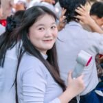 Seorang mahasiswi tersenyum sambil memegang kipas portable di tengah kerumunan pelajar