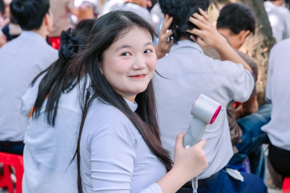 Seorang mahasiswi tersenyum sambil memegang kipas portable di tengah kerumunan pelajar