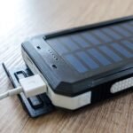 Powerbank tenaga surya hitam dengan kabel USB terhubung, diletakkan di atas meja kayu