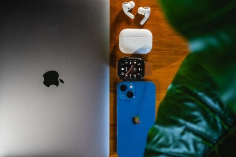Susunan laptop, earbud nirkabel, kotak pengisi daya, smartwatch, dan smartphone di atas meja kayu dengan daun hijau.