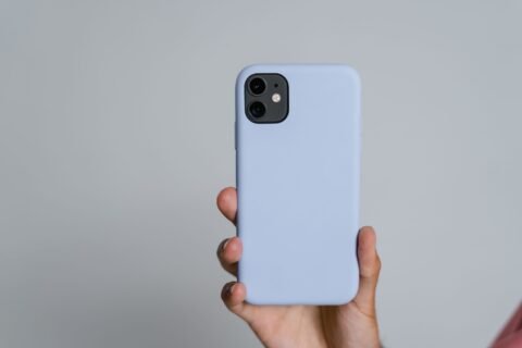 Seseorang memegang smartphone dengan casing polos berwarna biru muda di depan latar abu-abu.