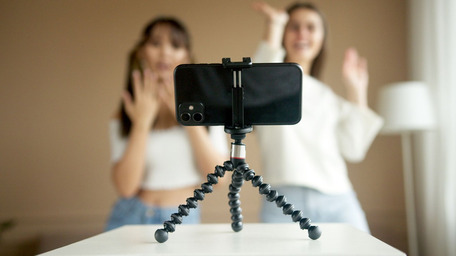 Smartphone terpasang pada tripod mini berkaki fleksibel di atas meja, dengan dua orang merekam video di latar belakang.