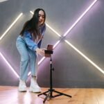 Perempuan mengatur smartphone yang terpasang pada tripod tinggi di ruangan dengan lampu neon.